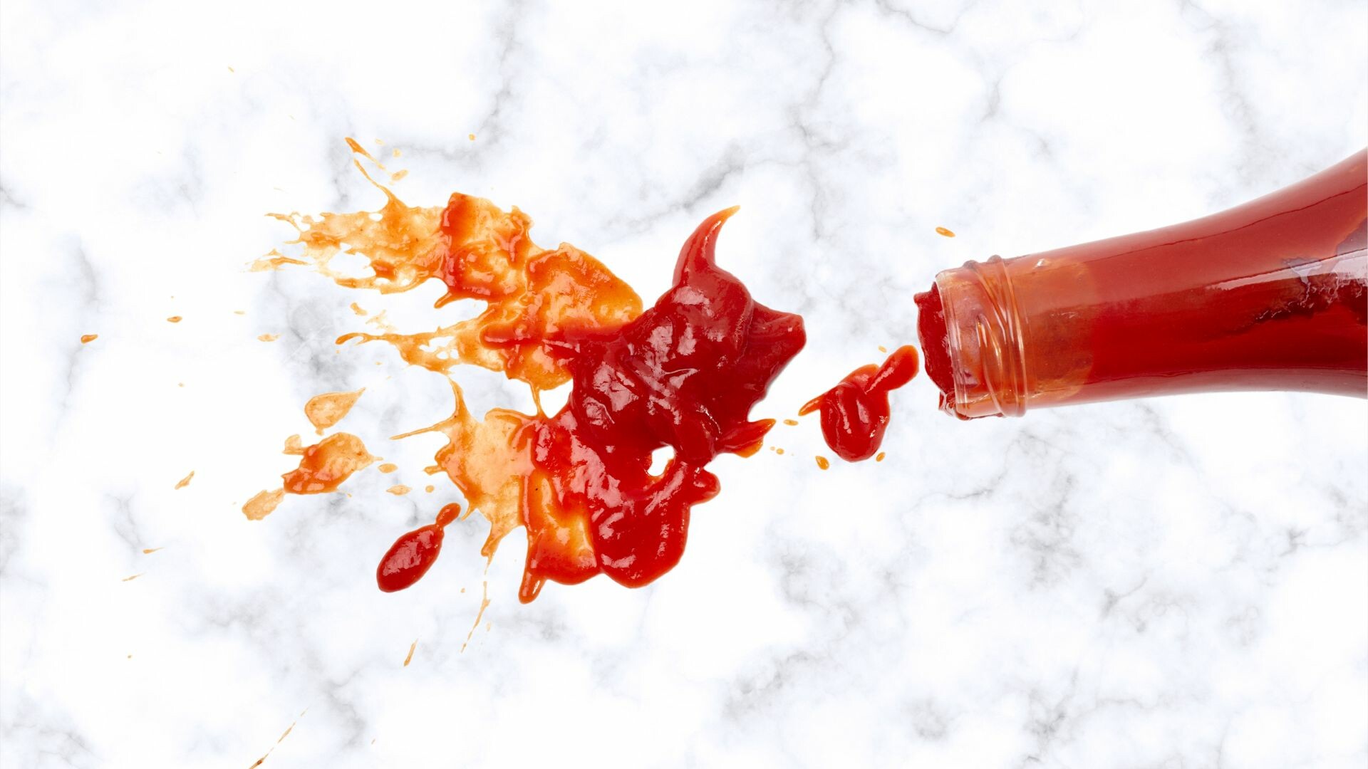 Glass bottle of ketchup that has spilled onto a surface.