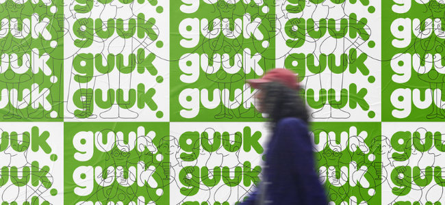 Person walking in front of wall of green and white posters with the words "guuk"